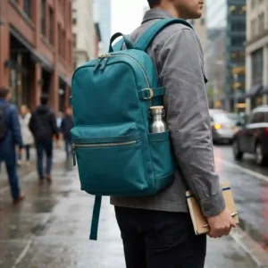 A commuter walking down a city street wearing a sleek teal backpack over a casual gray jacket.
