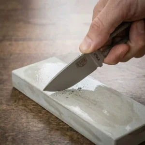 A person using a whetstone to sharpen the edge of a carbon steel fixed blade edc knife.